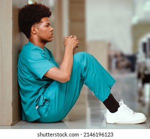 Emotional Stressed Intern Doctor Sitting Against Wall On Floor In Hospital Corridor. Distraught Medical Student In Seriously Depressed, Sad Or Exhausted. Overworked Male Nurse Sits In Despair Or Tired