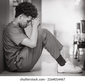 Emotional Stressed Young Doctor Sitting Against Wall On Floor In Hospital Corridor. Distraught Medical Student Depressed, Sad Or Exhausted. Overworked Black Man Nurse Sits In Despair. Black And White.