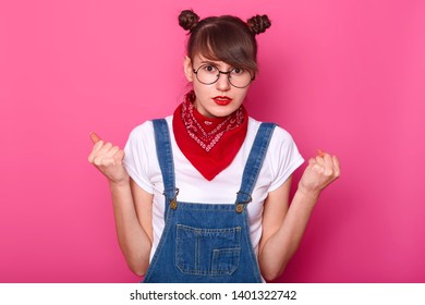 Emotional Student Clenches Fists, Cant Control Negative Emotions, Wears Round Spectacles, White T Shirt, Denim Overalls, Isolated Over Pink Studio Wall. People, Bad Feeling And Body Language Concept.