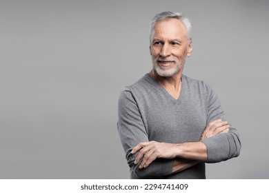 Emotional studio portrait of handsome confident elderly senior gray haired man in casual clothes, posing with his arms crosse, smiling looking away at copy advertising space, isolated grey background - Shutterstock ID 2294741409