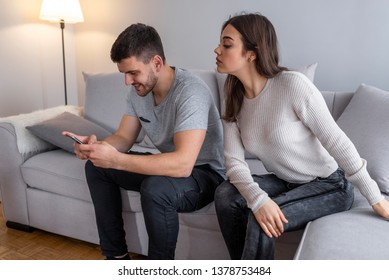 Emotional upset couple sitting on couch and quarreling about smartphone, mistrust concept. Jealous suspicious mad wife arguing with obsessed husband holding phone texting cheating on cellphone