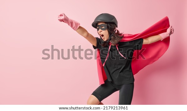 Emotional woman superhero has extraordinary or\
superhuman powers ready to do heroic deeds makes flying gesture\
wears helmet eyemask rubber gloves cloak isolated on pink\
background fights against\
evil