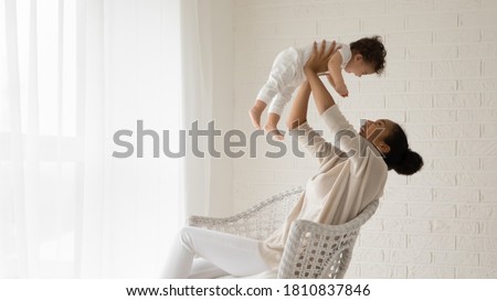 Emotional young african american mother sitting on comfortable chair, raising on straight arms laughing adorable toddler baby son or daughter, having fun playing together in light living room.