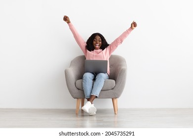 Emotional young african american woman in casual sitting in arm chair with laptop and raising hands up, happy black lady got dream job or great news, white empty wall background, copy space