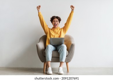 Emotional young Asian guy sitting in armchair with laptop, raising hands up in excitement, celebrating success against white studio wall, full length. Millennial man enjoying online sale or big win