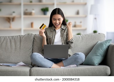 Emotional Young Asian Woman Sitting On Couch In Living Room And Using Laptop And Credit Card, Checking New Website For Online Shopping Or Banking, Home Interior, Copy Space, Catching Sales