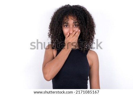 Emotional Young beautiful girl with afro hairstyle wearing black t-shirt over white background gasps from astonishment, covers opened mouth with palm, looks shocked at camera. Reaction concept