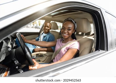 Emotional Young Black Family Husband And Wife Having Car Ride Together On Weekend, Looking At Camera And Smiling, Sun Flare. Happy Afro American Woman Driving Brand New White Automobile