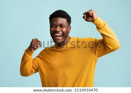 Emotional young black guy in casual raising fists up on blue studio background, celebrating success, happy handsome african american millennial man sharing good emotions, copy space