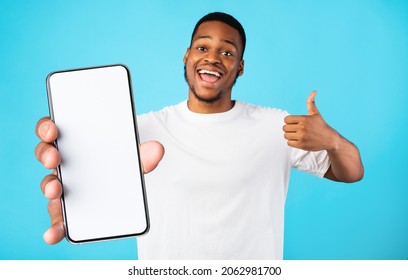Emotional young black man in white t-shirt demonstrating brand new smartphone with empty screen and showing thumb up, recommending newest mobile application, blue studio background, mockup