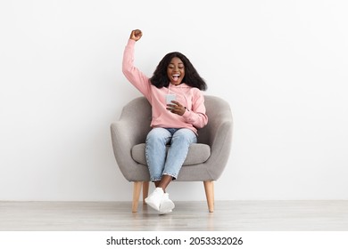 Emotional Young Black Woman Gambling Online Or Trading On Stocks And Markets, Celebrating Success, Raising Hand Up And Smiling, Using Cellphone While Resting In Arm Chair At Home, Copy Space