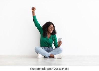 Emotional Young Black Woman Gambling Online Or Trading On Stocks And Markets, Celebrating Success, Raising Hand Up And Smiling, Using Cellphone While Resting Over White Background, Copy Space