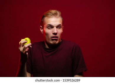 Emotional young blond man eating a green sour apple and posing against a dark red background