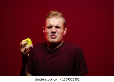 Emotional young blond man eating a green sour apple and posing against a dark red background