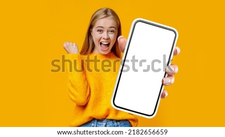 Emotional young blonde woman shouting and raising clenched fist in the air, rejoicing success over orange studio background, showing cell phone with white empty screen, gambling online, collage