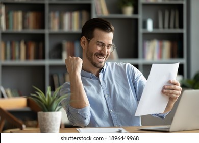 Emotional young businessman in eyeglasses reading paper notification, feeling excited of getting unbelievable news. Happy overjoyed man getting dream job offer, celebrating success in office.