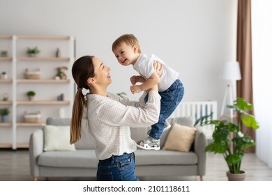 Emotional young caucasian mother raising laughing adorable toddler baby son, holding kid pretending flying, having fun and playing together in light living room, side view - Powered by Shutterstock