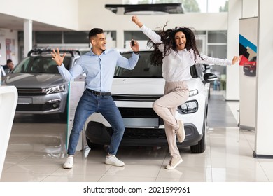 Emotional Young Couple Arab Guy And Curly Brunette Lady Dancing, Jumping And Screaming In Car Saloon, Holding Key From Their Brand New Auto, Celebrating Successful Purchasing, Full Length Photo