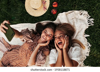 Emotional young tanned girls in stylish dresses take selfie and smile outdoors. Brunette woman holds phone. Charming ladies have picnic in park. - Powered by Shutterstock