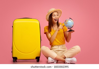 Emotional Young Woman With Globe Sitting Next To Yellow Luggage Over Pink Background, Pointing At New Destination To Travel, Copy Space. Excited Lady Getting Ready For Vacation, Choosing Country