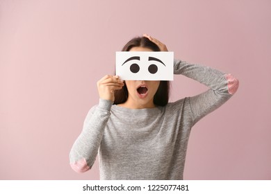 Emotional Young Woman Hiding Face Behind Sheet Of Paper With Drawn Eyes On Color Background