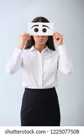 Emotional Young Woman Hiding Face Behind Sheet Of Paper With Drawn Eyes On Light Background