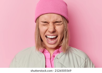 Emotional young woman screams loudly keeps mouth opened closes eyes feels annoyed wears hat and jacket poses against pink background. Irritated girl shouts agrily expresses negative emotions