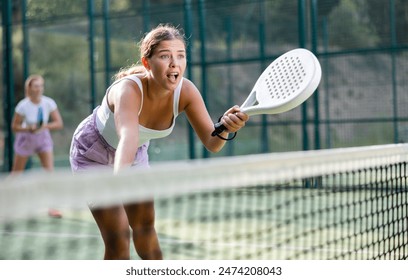 Emotional young woman tennis player with a padel racket training on an outdoor court, hitting the ball - Powered by Shutterstock