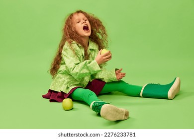 Emotionally eating fruits. Little cute girl, child with curly hair posing with apples over green studio background. Concept of childhood, emotions, fun, fashion, lifestyle, facial expression