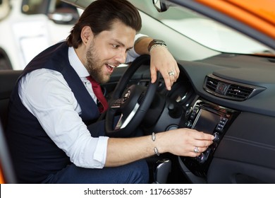 Emotionally Man In White Shirt And Vest Sitting In New Auto In Car Dealership. Male Customer Testing Vehicle Before Buying. Handsome Man Looking Down And Turned Radio In Car.