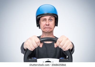 Emotionally Scared Man In Blue Motorcycle Helmet Looks At The Road With One Eye, Gripping The Steering Wheel In His Hands, On Light Background.