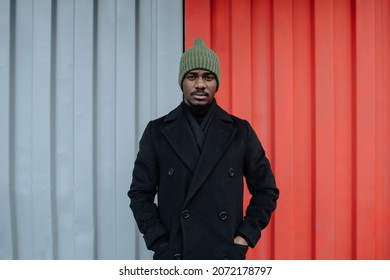 Emotionless black man in a warm autumn coat standing in front of a red and white corrugated sheet container wall