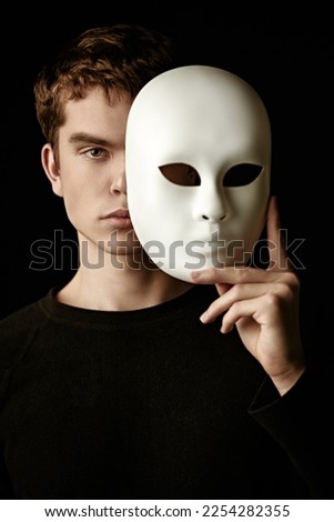 An emotionless young man in a black pullover holds a white mask in his hand, covering part of his face with it. Black studio background. Roles of people. Psychology, inner world.  Stock photo © 