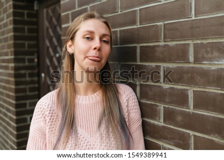 Emotions. Cute young blonde woman shows tongue. In the background, a brick wall