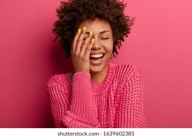 Emotions, Lifestyle Concept. Overjoyed Dark Skinned Woman Laughs With Closed Eyes, Hears Good Joke, Covers Face With Palm, Has Nice Conversation, Chuckles And Shows White Teeth, Wears Knitted Sweater