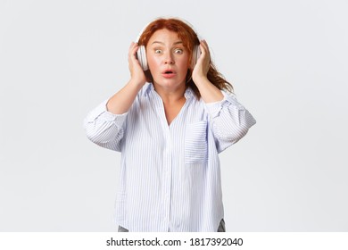 Emotions, lifestyle and leisure concept. Surprised and amazed redhead adult woman react to song or music in headphones, stare astonished at camera, standing white background - Powered by Shutterstock