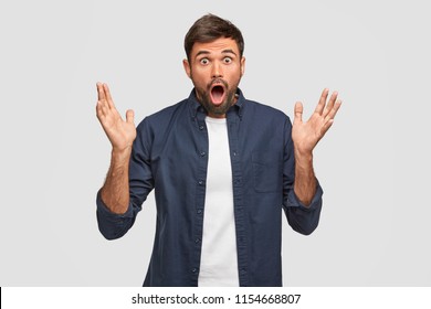 Emotive bearded European male with amazed expression, gestures with hands, keeps palms raised, reacts on sudden news, keeps jaw dropped, poses against white background. Surprisement concept.