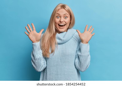 Emotive blonde young woman raises hands and has happy face expression feels excited dressed in knitted jumper isolated over blue background. Impressed European female being full of happiness.