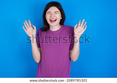 Emotive Caucasian woman wearing purple T-shirt isolated over blue background laughs loudly, hears funny joke or story, raises palms with satisfaction, being overjoyed amused by friend
