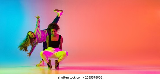 Emotive dance style. Portrait of two young beautiful hip-hop grils dancing on colorful gradient blue orange background in neon. Youth culture, movement, active lifestyle, action, street dance, ad