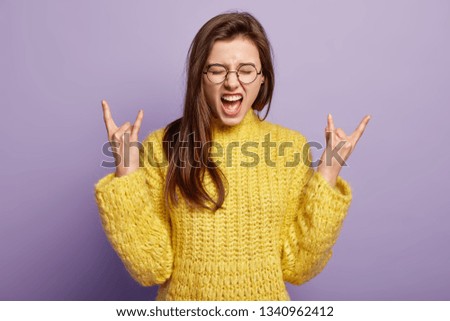 Emotive European woman makes horns with fingers, raises hands, exclaims loudly, wears yellow jumper, poses over purple background. Impressed lady shows rock n roll gesture. I like heavy metal Foto stock © 
