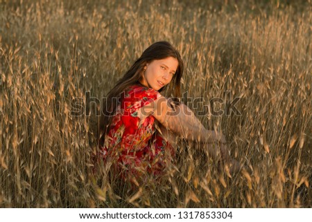 Emotive portrait of a happy and slim beautiful teenager girl looking into the camera while sitting in a field of wheat lit by the rays of the setting sun. Summertime. Summer vacation.Positive emotions