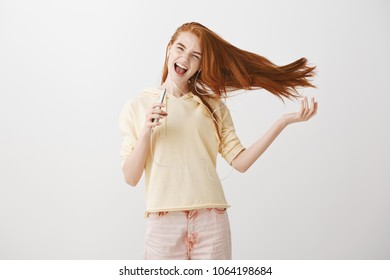 Emotive Woman With Powerful Voice Thrilled To Goosebumps With Great Sound Of Earphones. Cheerful Attractive Redhead Holding Smartphone Like Microphone, Listening Music, Singing Along While In Earbuds