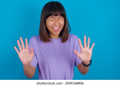 Emotive Young beautiful asian girl wearing purple t-shirt over blue background laughs loudly, hears funny joke or story, raises palms with satisfaction, being overjoyed amused by friend