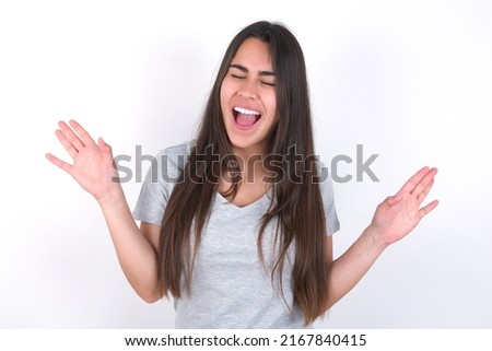 Emotive young caucasian woman wearing grey t-shirt over white background laughs loudly, hears funny joke or story, raises palms with satisfaction, being overjoyed amused by friend
