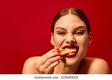 Emotive young woman eating fried potato, fries over vivid red background. Junk food lover. Delicious taste. Food pop art photography. Modern style. Complementary colors. Copy space for ad, text
