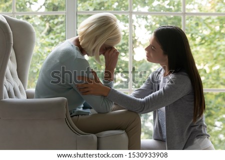 Empathic millennial lady soothing desperate crying mature mother, sitting in armchair. Side view upset grown up daughter comforting, supporting caring unhappy middle aged mommy felling unwell at home.