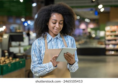 An employee with afro hair, wearing an apron, uses a tablet in a grocery store for inventory or customer service. This showcases efficient and friendly customer interaction in a modern market - Powered by Shutterstock