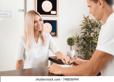 The Employee Of The Beauty Salon Meets The Client In The Reception Of A Modern Beauty Salon. A Man Signs A Paper With The Consent For Maintenance. The Woman Smiles At Him.