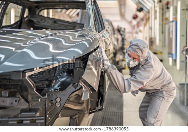 An employee of the car body paint\
shop in white gloves and a medical mask on his face checks the\
quality of the painted surface. Working during the\
pandemic